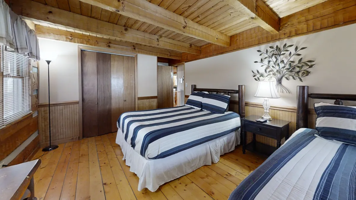 This image features two beds with striped bedding, decorative wall art, a nightstand with a lamp, and wooden flooring. The inviting and relaxed mood hints at a welcoming space ideal for both resting and light socializing, perfectly suited to families or groups.