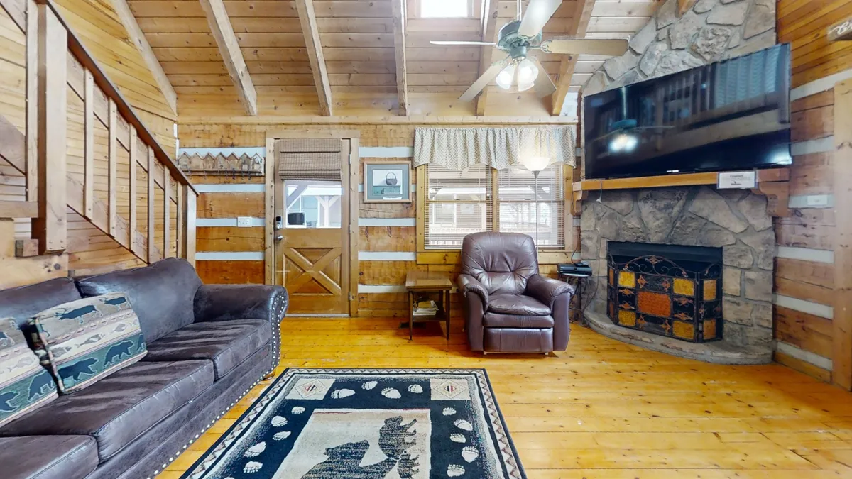 A rustic living room centered around a striking stone fireplace, with a large TV, leather recliner, and exposed wooden beams. The arrangement provides a cozy environment for relaxing by the fire or enjoying entertainment with friends and family.