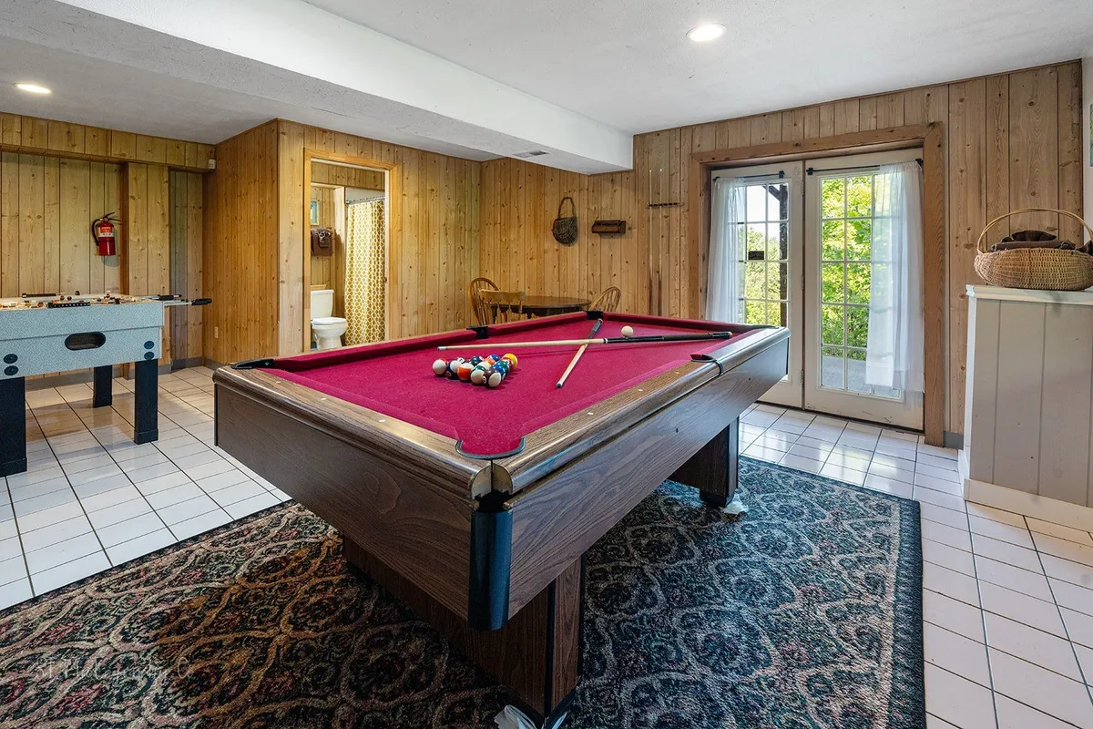 A spacious game room featuring a full-size pool table and a foosball table set against rustic wood-paneled walls. Double doors in the background suggest convenient access to the outdoors, while casual seating and warm lighting create an inviting atmosphere ideal for socializing and play.