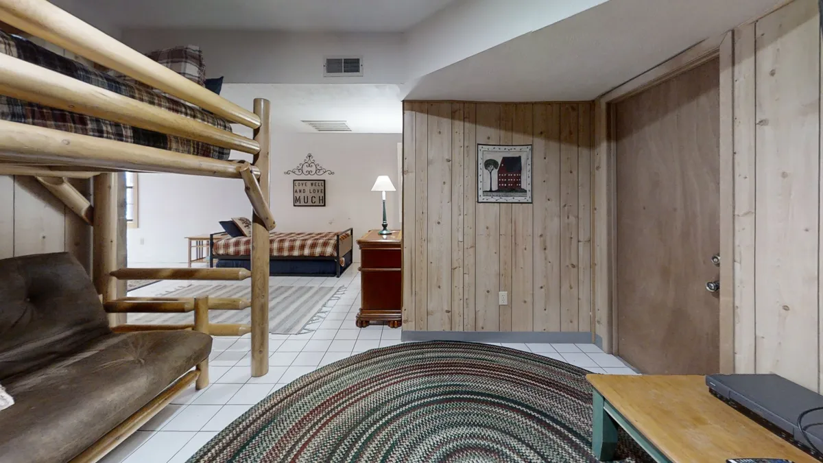 A cozy room featuring a bunk bed and a sofa, set against wood-paneled walls and highlighted by soft lighting and playful artwork. The rug and lampshade add warmth, suggesting a relaxed, social space ideal for friends to unwind and connect.