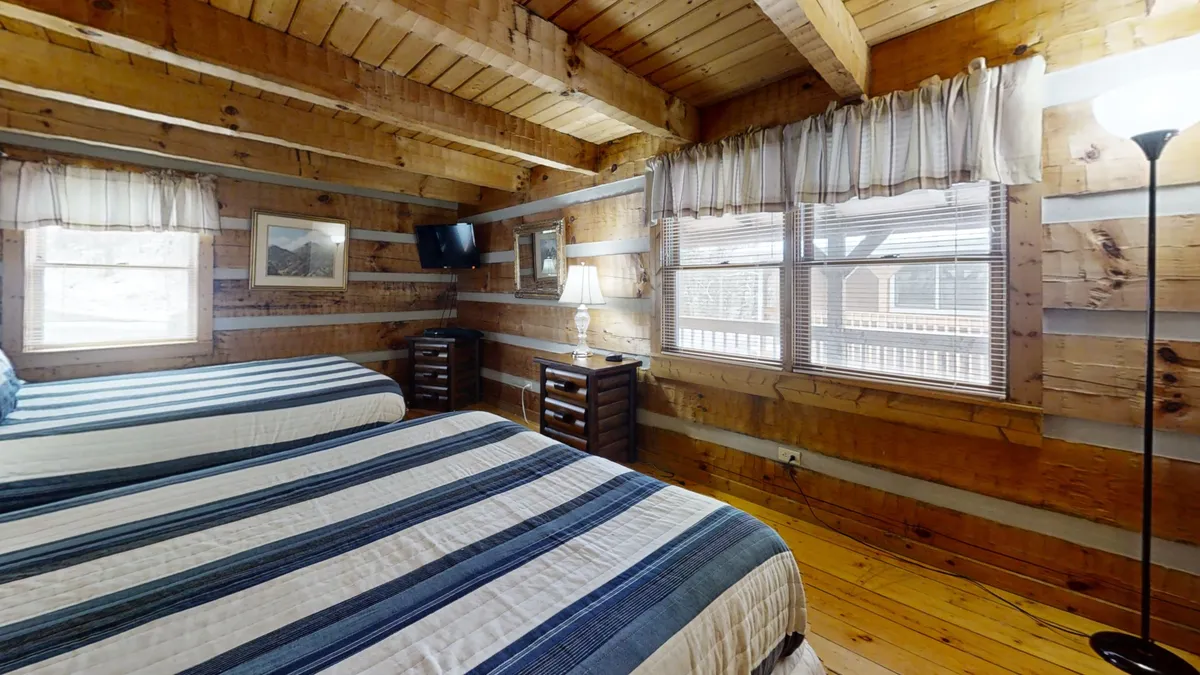 A warm, inviting bedroom featuring two beds with striped bedding, wooden beams and walls, and large windows with curtains that let in natural light. There's a cozy table lamp between the beds and a reading nook by the window, suggesting space for multiple family members to relax, converse, or unwind together.
