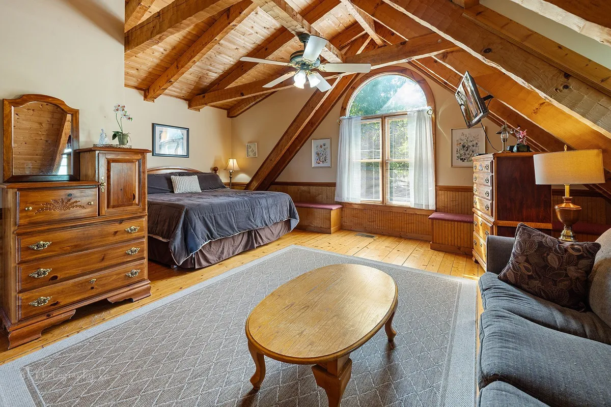 This image captures a spacious, inviting common area with sturdy wooden beams, a large window that floods the room with natural light, a comfortable sofa, and a cozy bed. The scene hints at guests relaxing by the window, enjoying the serene atmosphere while sipping morning coffee or reading. The blend of comfort and scenic views is apparent, making it a welcoming hub for group gatherings.