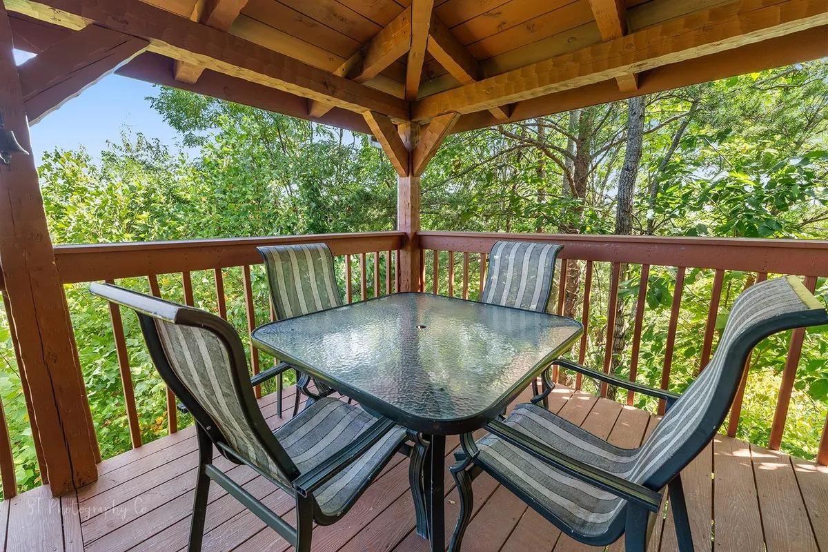An exterior deck scene with a table and chairs overlooking greenery, framed by a wooden railing. The setting is ideal for outdoor dining, morning coffee, or simply soaking in the natural surroundings.
