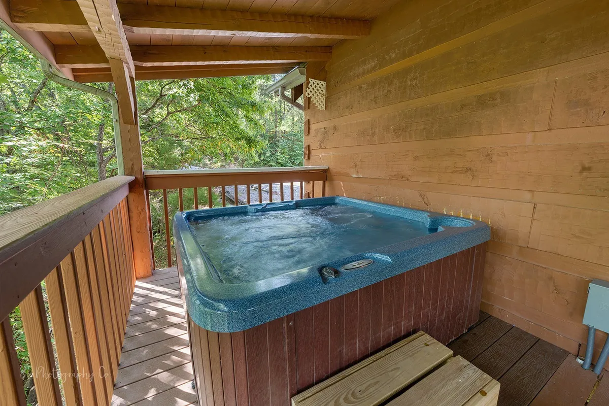 A bubbling hot tub sits on a wooden deck, surrounded by lush greenery. The setting evokes relaxation, with the hot tub inviting you to soak while enjoying the natural mountain environment. The open-air arrangement hints at mountain breezes and starlit skies.
