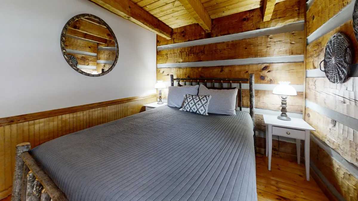 A rustic bedroom with a wooden bed frame, decorative pillows, round mirror, and wood-paneled walls. The mood is inviting and warm, ideal for relaxing, reading, or quiet conversation, with a serene ambiance that suggests restful nights after busy days of exploring the Smokies.