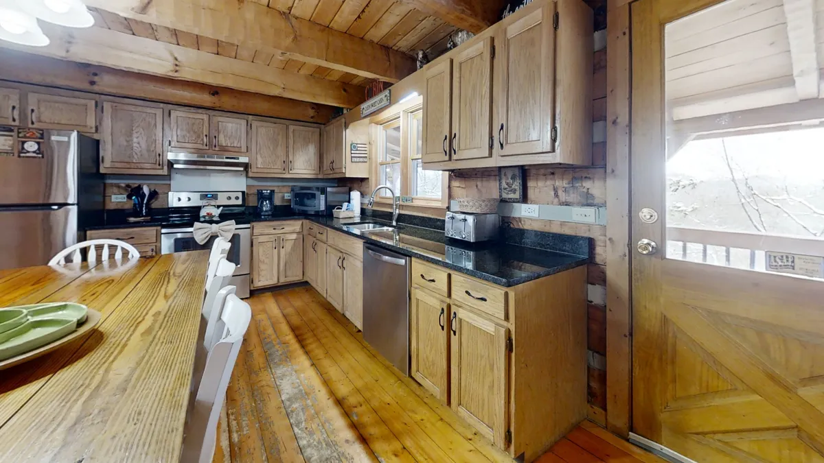 A spacious wooden dining area with a large table set near a window that bathes the space in natural light. The adjacent kitchen with black granite countertops and modern appliances is visible, inviting group gatherings for meals and friendly conversation.