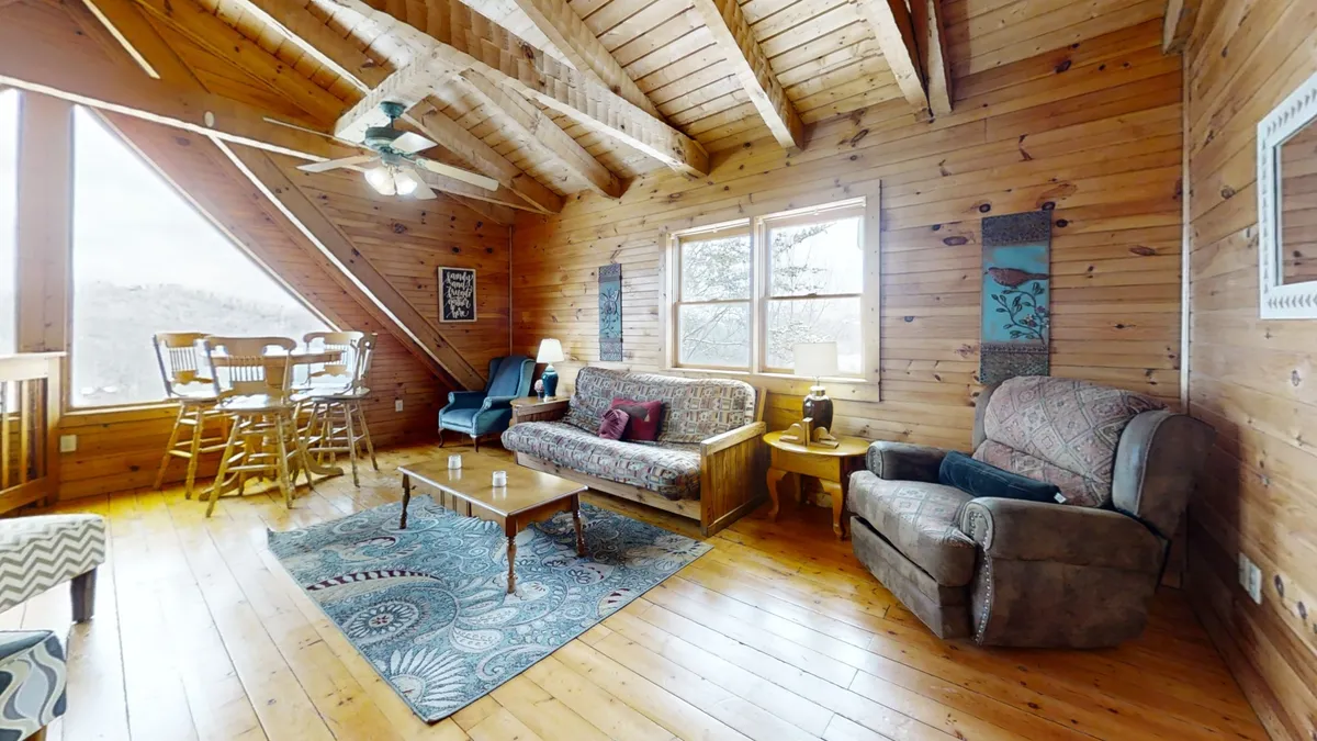 This image features a comfortable, open-plan living and dining area with large windows that let in natural light and provide views of the surrounding landscape. Visible are wooden beams, a cozy sofa with decorative pillows, and a dining area with high chairs, all set in a space designed for both relaxation and entertaining.