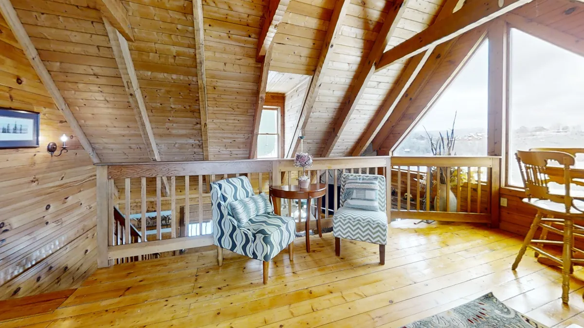 An airy loft space with wooden beams, cozy chairs, a small table for board games, and large windows offering panoramic mountain views. The room is bathed in natural light, creating a warm, relaxing retreat above the main level.