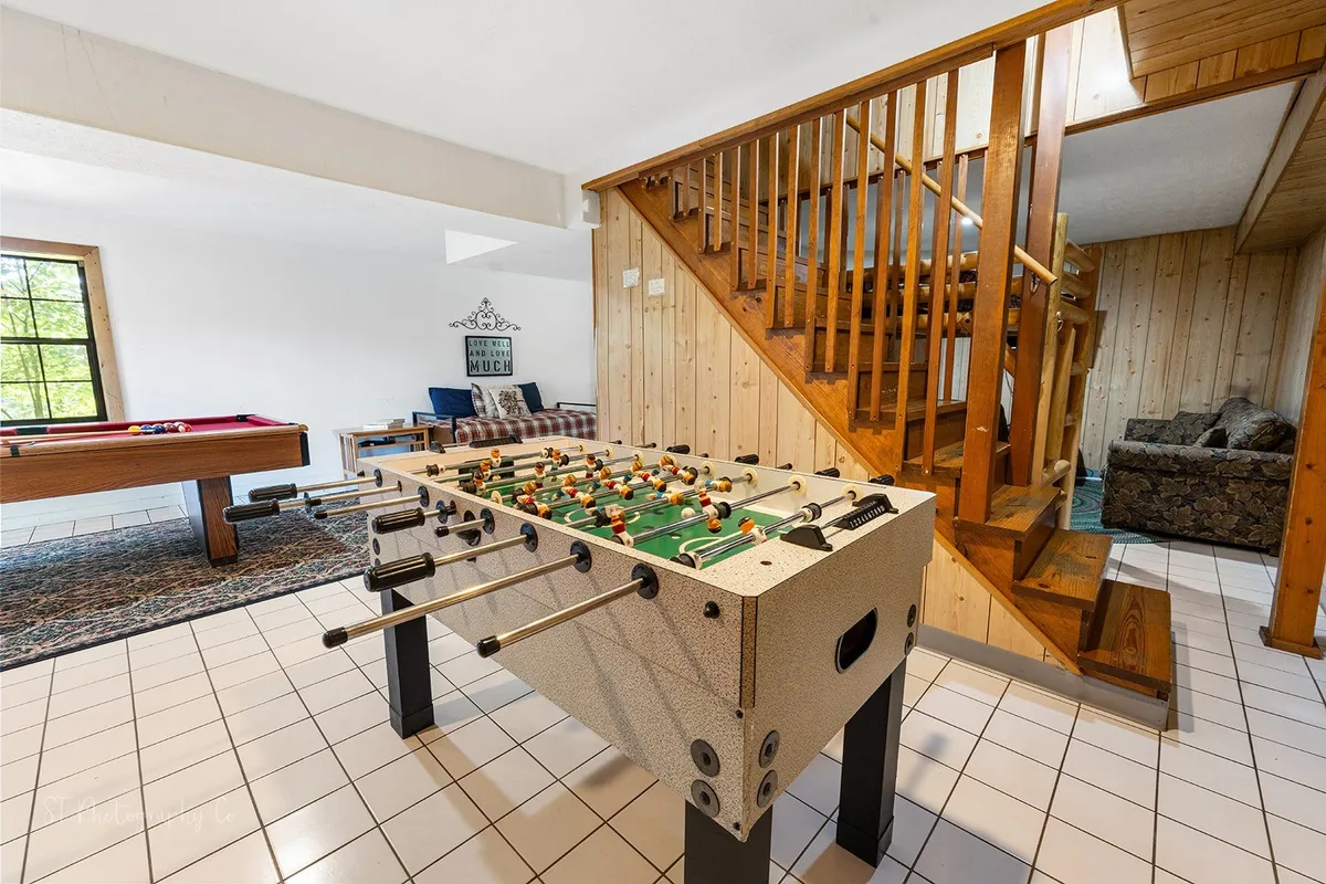 A bright, playful room with a foosball table, pool table, and a comfortable couch for hanging out. A wooden staircase leads upstairs, hinting at the transition to the loft, while natural light floods the space, making it warm and inviting.
